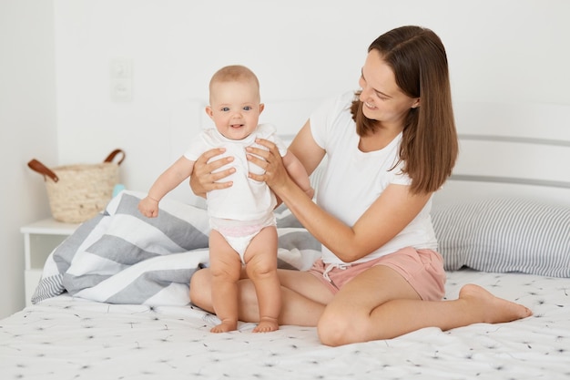 Portret Pozytywnej Zadowolonej Kobiety Ubrana W Białą Koszulę I Krótkie Siedzenie Na łóżku Z Córką W Kolorze Ciała Matka Trzyma Jej Stojącego Malucha Dziecko Ciemnowłosa Kobieta Bawi Się Z Dzieckiem