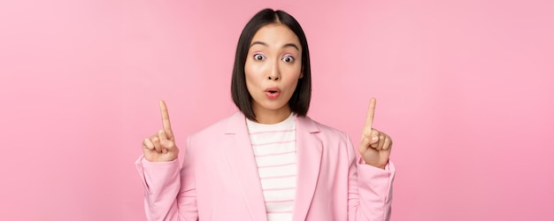 Portret pracownika korporacyjnego azjatyckiej bizneswoman w garniturze wygląda na zaskoczonego punktami reklamowymi, wskazującymi na baner lub logo na szczytach stoisk na różowym tle