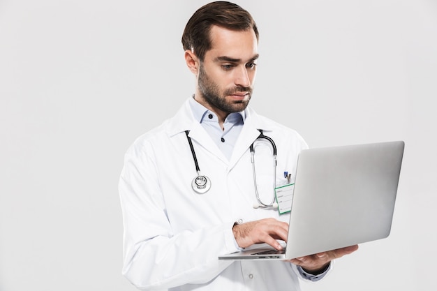 Portret profesjonalnego młodego lekarza ze stetoskopem pracującym w klinice i trzymającego laptopa na białym tle nad białą ścianą