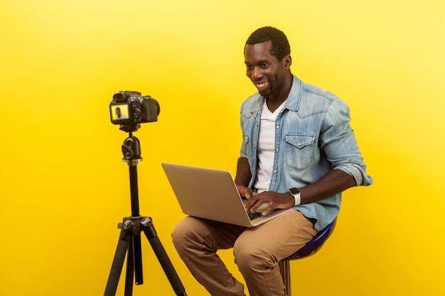 Portret profesjonalnego wesołego fotografa w dżinsowej koszuli komponującej scenę fotograficzną na laptopie podczas robienia zdjęcia cyfrowym aparatem dslr. strzał w studio na białym tle na żółtym tle