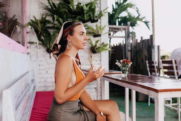 Portret profil atrakcyjna ładna dziewczyna w letnie ubrania siedzi w stylowej restauracji na świeżym powietrzu z kawą z egzotycznymi roślinami