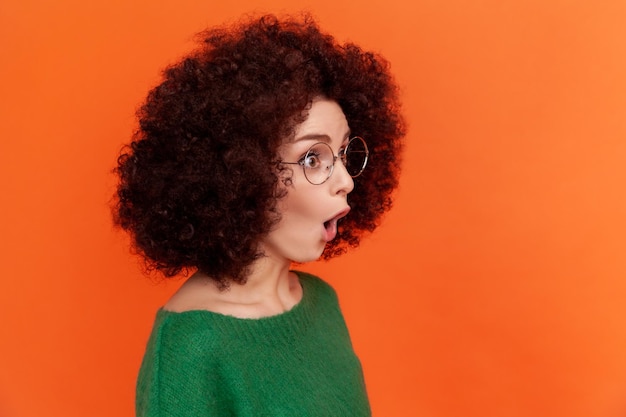 Portret profil zszokowana kobieta z fryzurą afro w zielonym stylu casual sweter stojący z otwartymi ustami, odwracając wzrok widzi coś zaskakującego Kryty studio strzał na białym tle na pomarańczowym tle