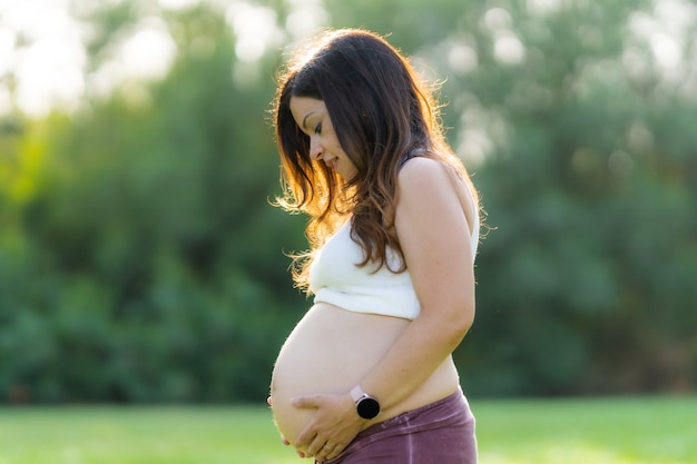 Portret profilowy dorosłej kobiety w ciąży latynoskiej dotykającej jej brzucha podczas noszenia wygodnej odzieży sportowej z brzuchem w powietrzu w parku z promieniami słonecznymi świecącymi na nią