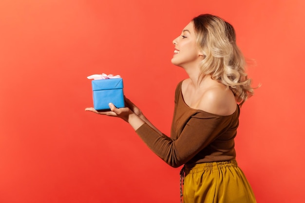 Portret profilowy szczęśliwa kobieta trzymająca na ręku niebieskie pudełko, dające prezent i uśmiechnięty Kryty studio strzał na białym tle na pomarańczowym tle