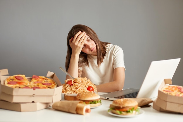 Portret przepracowanej kobiety pracującej na laptopie siedzącej w otoczeniu dużej różnorodności fast foodów trzyma dłoń na czole odizolowanym na szarym tle