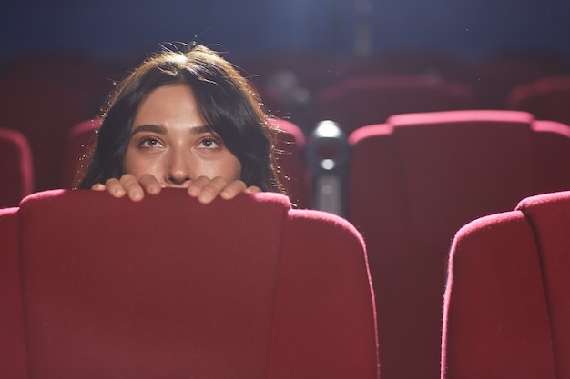 Portret przerażonej młodej kobiety chowającej się za siedzeniem podczas oglądania horroru w pustej sali kinowej, kopia przestrzeń