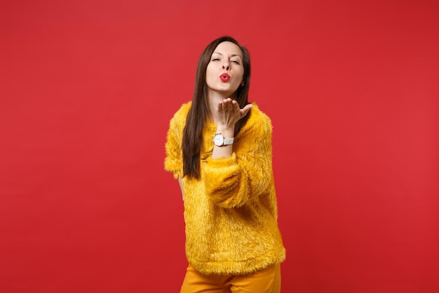 Portret przetargu całkiem młoda kobieta w żółtym futrze sweter dmuchanie, wysyłając pocałunek powietrza na białym tle na tle jasnej czerwonej ściany w studio. Ludzie szczere emocje, koncepcja stylu życia. Makieta miejsca na kopię.