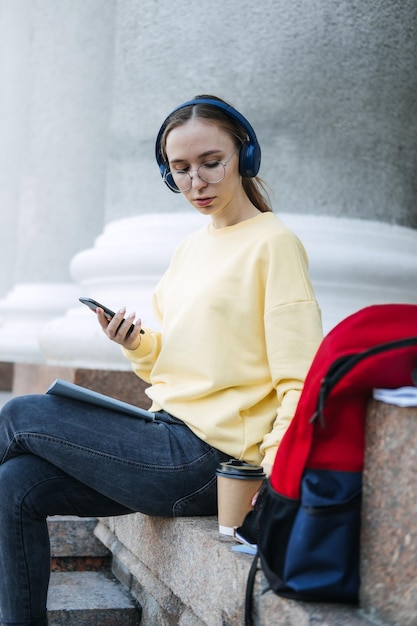 Portret przypadkowej studentki w słuchawkach przy użyciu aplikacji na telefon komórkowy na zewnątrz młoda kobieta studentka
