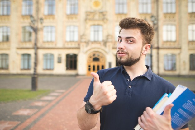 Portret Przystojna Studencka Pozycja Z Książkami W Rękach Uniwersyteckiego Budynku Tło I Pokazywać Aprobaty.