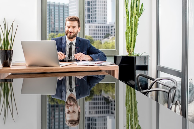 Portret przystojnego bankiera pracującego z laptopem siedzącym w luksusowym wnętrzu biurowym z pięknym widokiem na drapacze chmur