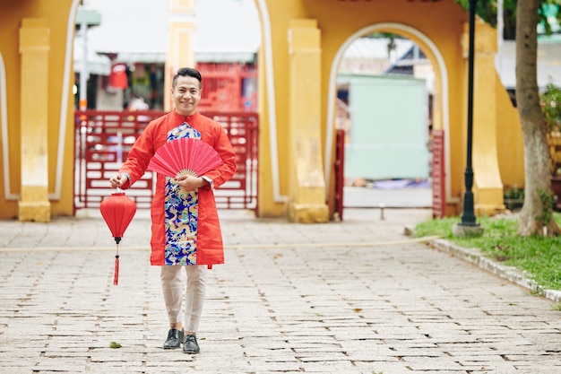 Portret przystojnego młodego azjatyckiego mężczyzny w tradycyjnym stroju na chiński nowy rok, stojącego na zewnątrz z wachlarzem papieru i czerwoną latarnią