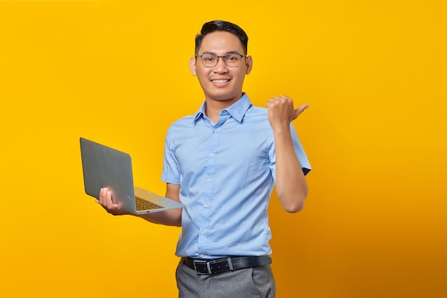 Portret przystojnego uśmiechniętego azjatyckiego mężczyzny w okularach za pomocą laptopa i wskazując palcami na miejsce na kopii na żółtym tle koncepcja biznesmena i przedsiębiorcy