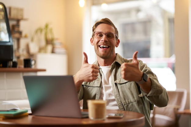 Portret przystojnego uśmiechniętego mężczyzny z podniesionymi kciukami siedzi w kawiarni z filiżanką kawy i laptopem zatwierdza