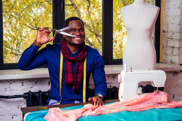 Portret przystojny afrykański mężczyzna uśmiechający się krawcowa z maszyną do szycia. Afrio amerykański mężczyzna stylowy projektant pracujący w manekinie warsztat krawiecki, taśma pomiarowa stołu w pokoju przed jesiennym oknem