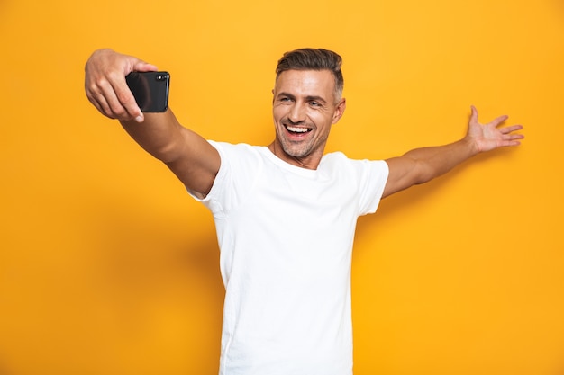 Portret przystojny mężczyzna w wieku 30 lat w białej koszulce śmiejącej się i robiący selfie na telefonie komórkowym odizolowanym na żółto