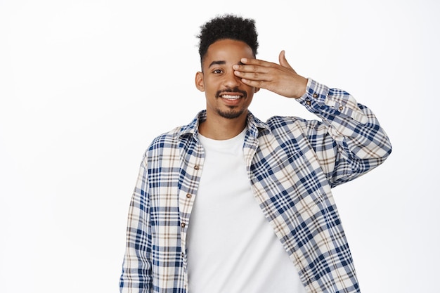 Portret przystojny młody afroamerykanin zakrywa jedno oko, uśmiechając się szczęśliwy i zrelaksowany, patrząc na kamerę, sprawdzając wzrok, koncepcję sklepu z okularami, białe tło