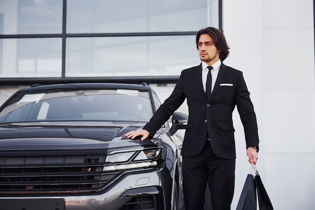 Portret przystojny młody biznesmen w czarnym garniturze i krawat na zewnątrz w pobliżu nowoczesnego samochodu i torby na zakupy.