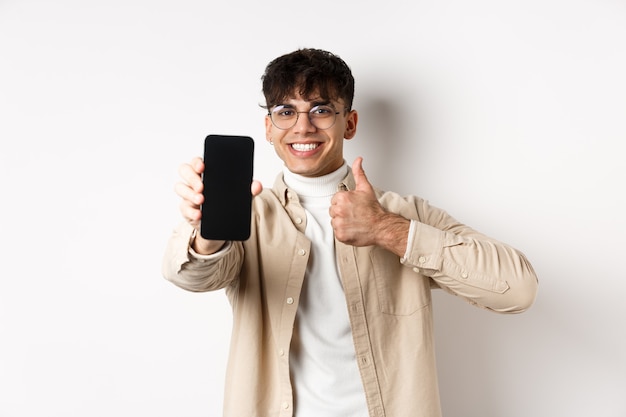 Portret przystojny młody mężczyzna w okularach, pokazujący pusty ekran smartfona i kciuk w górę, polecający aplikację internetową lub sklep, stojący zadowolony na białej ścianie.