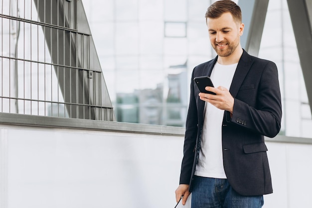 Portret przystojny nowoczesny mężczyzna biznesmen w garniturze spacerujący, uśmiechający się i czytający lub piszący wiadomość na telefonie na tle miejskich budynków i biur