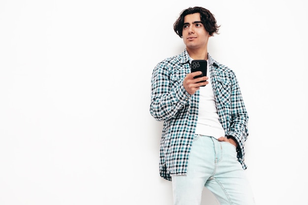 Portret przystojny pewnie model seksowny stylowy mężczyzna ubrany w kraciastą koszulę i dżinsy Moda hipster mężczyzna pozowanie w pobliżu białej ściany w studio trzymając smartfon za pomocą aplikacji na telefon komórkowy