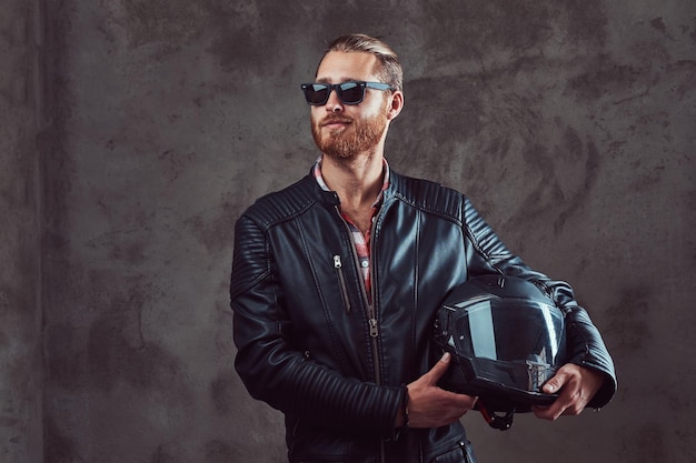 Portret przystojny pewnie stylowe rude rowerzysta w czarnej skórzanej kurtce i okularach przeciwsłonecznych, posiada kask motocyklowy, pozowanie w studio. Na białym tle na ciemnym tle.