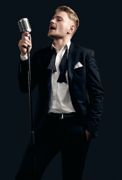 Portret przystojny piosenkarz blondyn w eleganckim smokingu i muszce z rocznika mikrofonem na czarnej ścianie
