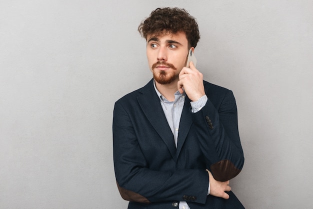 portret przystojny poważny młody biznesmen na białym tle nad szarą ścianą rozmawia przez telefon komórkowy.