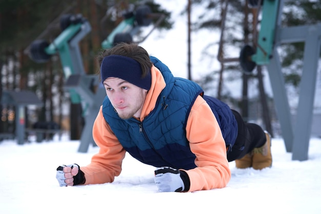 Portret przystojny sprawny silny lekkoatletycznego faceta, młody sportowy mężczyzna jest oszalowanie, robienie ćwiczeń poza deski, trening fitness w siłowni na świeżym powietrzu w zimę śnieżną pogodę, leżąc na śniegu. Zdrowy tryb życia