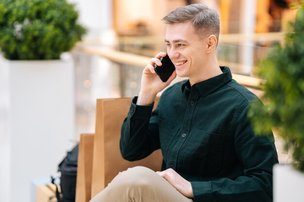 Portret przystojny uśmiechnięty młody mężczyzna rozmawia przez telefon komórkowy, siedząc na ławce w centrum handlowym