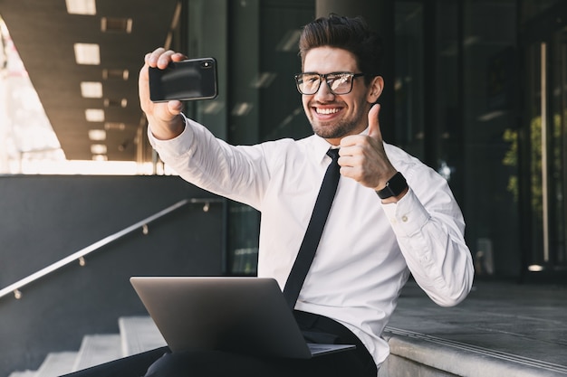 Portret radosnego biznesmena ubranego w formalny garnitur, siedzącego na zewnątrz szklanego budynku z laptopem i robienie zdjęć selfie na smartfonie