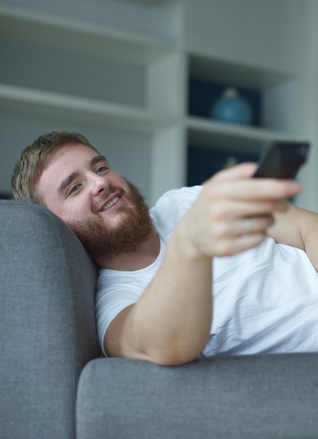 Portret radosnego młodego mężczyzny w przypadkowych ubraniach siedzącego i leżącego na kanapie w salonie i patrzącego na telewizor