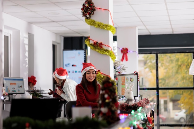 Portret radosnej azjatyckiej bizneswoman pracującej w okresie świątecznym w urządzonym biurze. Uśmiechnięta pracownica rozwiązująca zadanie przy biurku w różnorodnym miejscu pracy podczas zimowych świąt