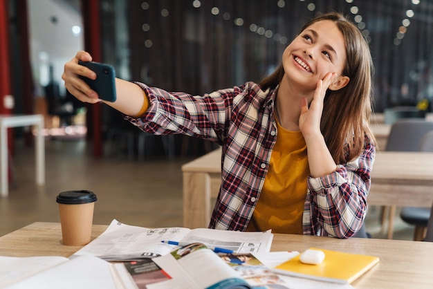 Portret radosnej dziewczyny biorącej selfie na telefon komórkowy i uśmiechającej się podczas odrabiania lekcji w bibliotece college'u