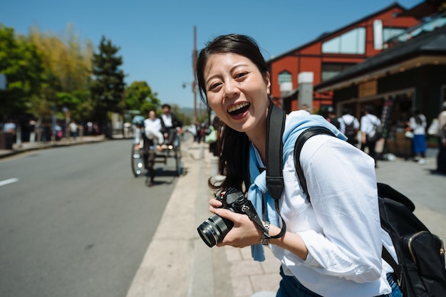 portret radosnej tajwańskiej dziewczyny trzymającej lustrzankę patrzy w kamerę z wielkim uśmiechem na chodniku głównej ulicy Arashiyama w japonii z rikszą w tle