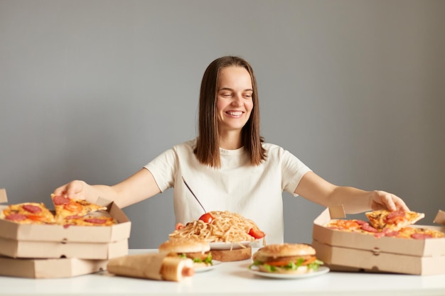 Portret radosnej, uroczej kobiety o brązowych włosach w białej koszulce, siedzącej przy stole wśród różnych fast foodów, biorącej pizzę z dwóch pudełek w tym samym czasie, pozując na białym tle na szarym tle