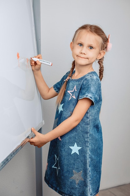 Portret radosnej uśmiechniętej ładnej dziewczyny stojącej przed dużą białą tablicą i rysującej na niej pomarańczowym markerem. Mała koncepcja artysty. zdjęcie z szumem