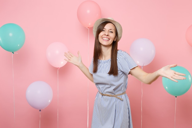 Portret radosny piękna młoda kobieta nosi słomkowy letni kapelusz i niebieską sukienkę rozkładając ręce na pastelowym różowym tle z kolorowymi balonami. Urodziny wakacje party ludzie szczere emocje.