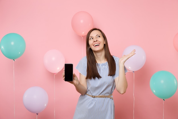 Portret radosny roześmiany młoda kobieta ubrana w niebieską sukienkę trzymającą telefon komórkowy z pustym pustym ekranem patrząc na pastelowe różowe tło z kolorowymi balonami. Koncepcja strony urodziny wakacje.