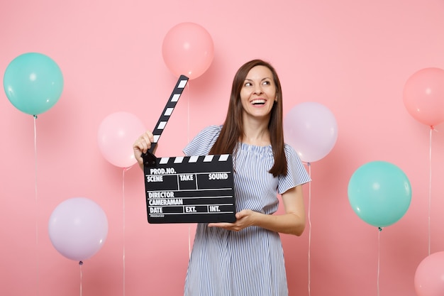 Portret roześmiana młoda kobieta w niebieskiej sukience patrząc w górę trzymając klasyczny czarny film co clapperboard na różowym tle z kolorowymi balonami. Urodziny wakacje, ludzie szczere emocje.