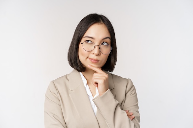 Portret rozważny azjatykci bizneswoman w szkłach robi założenie myślący stojący w beżowym kostiumu przeciw białemu tłu