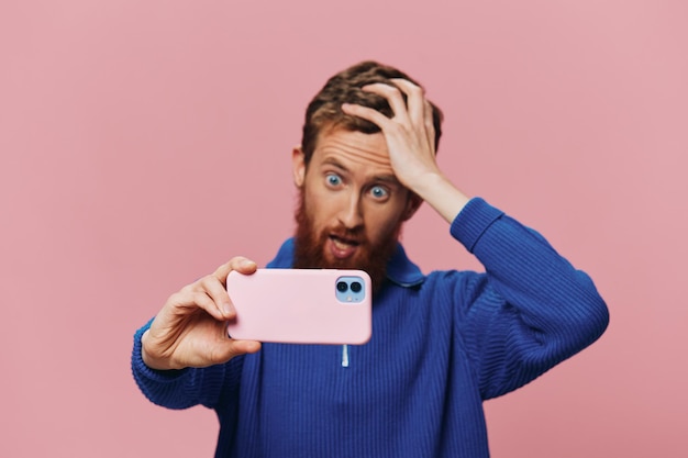 Portret rudego mężczyzny z telefonem w ręku robiącego selfie i zdjęcia na telefonie z uśmiechem na różowym tle blogera