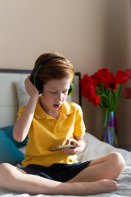 Portret rudowłosego chłopca dziecko słucha muzyki w słuchawkach siedzi na łóżku trzyma telefon w dłoniach pionowe zdjęcie