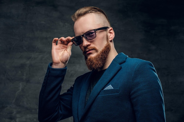 Portret rudy brodaty mężczyzna hipster ubrany w niebieską kurtkę i okulary przeciwsłoneczne na szarym tle.