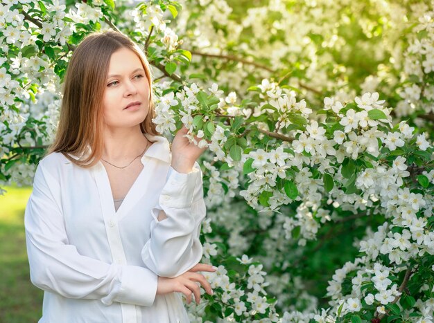 Portret samotnej młodej damy z przemyślanym uśmiechem i białą bluzką stojącej wśród kwitnących drzew