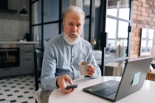 Portret siwowłosego dojrzałego starszego biznesmena pracującego na laptopie i używającego smartfona trzymającego filiżankę kawy siedzącego przy stole w pokoju kuchennym rano patrzącego na kamerę