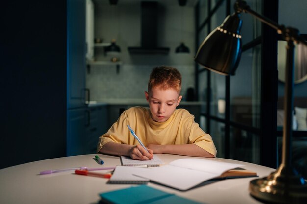 Portret skoncentrowanego ucznia uczącego się w domu, odrabiającego lekcje, siedzącego przy stole pod światłem lampy wieczorem Uczeń piszący w zeszycie w nocy