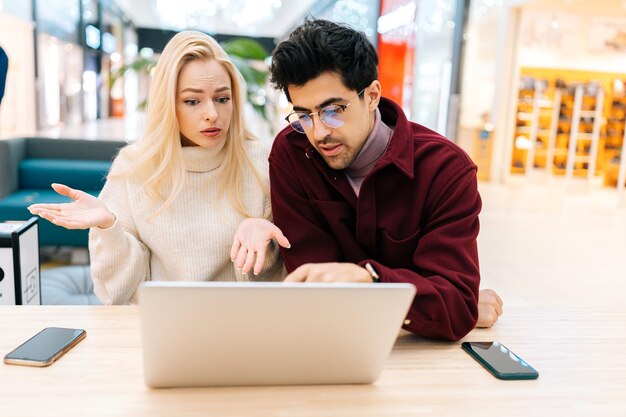 Portret skoncentrowanej młodej pary używającej laptopa do pisania razem, omawiającej zakup w Internecie, patrzącej na ekran siedzący przy stole w holu