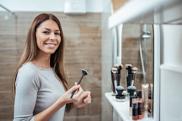 Portret śliczna milenijna kobieta z makijażu muśnięciem w łazience w domu, patrzeje kamerę.