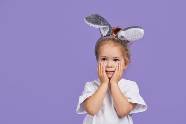 Portret śliczna uśmiechnięta małe dziecko dziewczyna jest ubranym królików ucho na Wielkanocnym dniu. Śmieszne emocje na fioletowym na białym tle miejsca