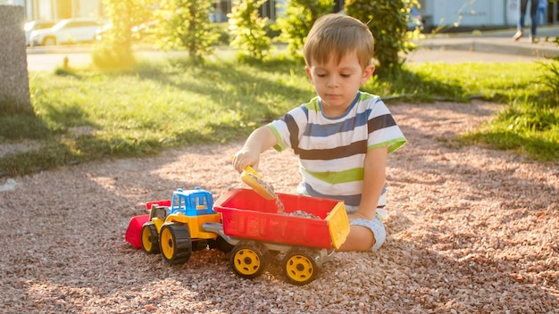 Portret ślicznego 3-letniego chłopca siedzącego na placu zabaw w parku i bawiącego się kolorową ciężarówką z tworzywa sztucznego